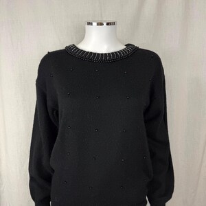 May include: Black sweater with a round neckline and long sleeves. The neckline is trimmed with a beaded design, and small beads are scattered across the front. The sweater is displayed on a mannequin.