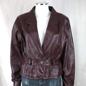May include: A burgundy leather jacket with a notched collar, button closure, and a belted waist. The jacket has long sleeves and is displayed on a mannequin. The jacket is a classic style.