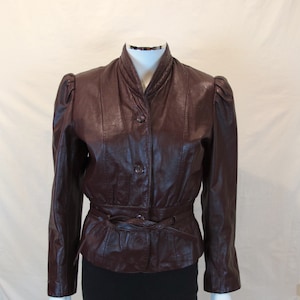May include: A brown leather jacket with a belted waist and a mandarin collar. The jacket has a button closure and long sleeves.