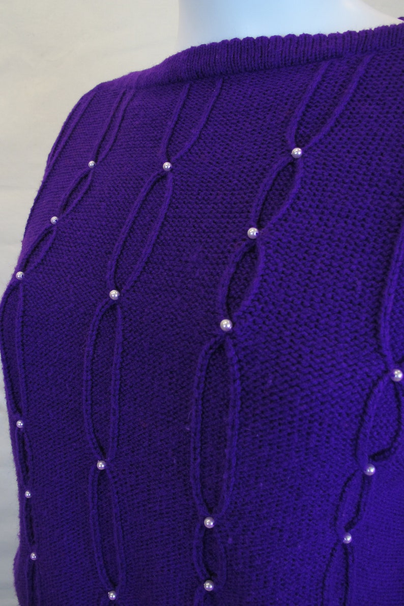 Puede incluir: Un su&eacute;ter de punto morado con un patr&oacute;n de punto de cable y adornos de perlas.