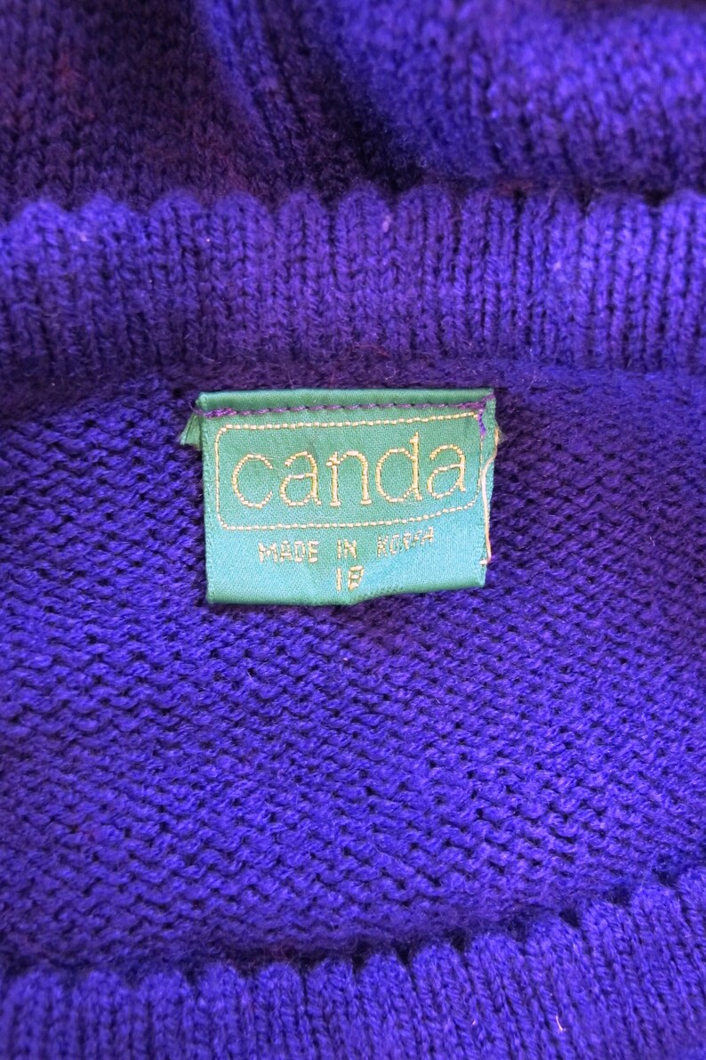 Puede incluir: Un su&eacute;ter de punto morado con una etiqueta verde que dice "canda" y "made in korea" en texto blanco.