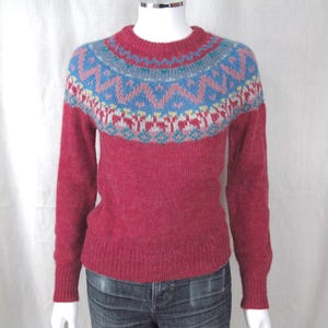 May include: A red sweater with a patterned yoke in blue, pink, and yellow. The sweater has long sleeves and a crew neck. The garment is displayed on a mannequin.