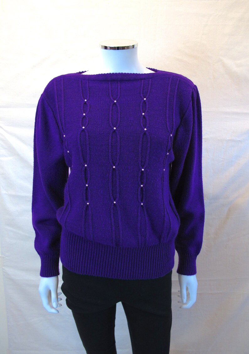 Puede incluir: Un su&eacute;ter de punto morado con un dise&ntilde;o de punto de cable y adornos de perlas. El su&eacute;ter tiene mangas largas y un cuello redondo.