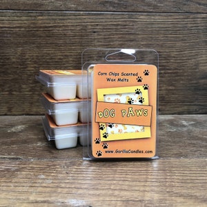 May include: A stack of six white wax melts in a clear plastic container. The container has a label that says "Corn Chips Scented Wax Melts" and "DOG PAWS" with a paw print design. The label also includes the website "www.GorillaCandles.com".
