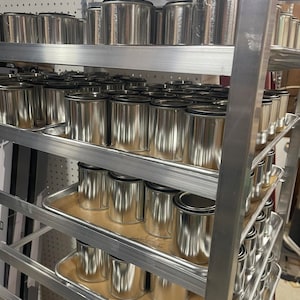 May include: A metal shelving unit with multiple shelves filled with silver paint cans. The shelves are made of metal and have a grid pattern. The cans are all the same size and shape, and they are arranged in rows. The cans are labeled with the word "Dirt".