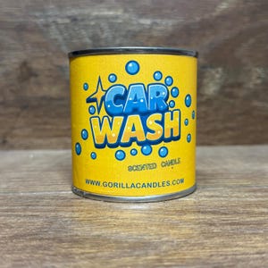 May include: A yellow scented candle in a metal container. The candle has the words "CAR WASH" in blue and white, surrounded by blue bubbles. The website www.gorillacandles.com is printed at the bottom.