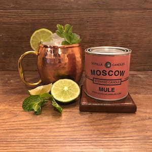Puede incluir: Un mug de cobre para Moscow Mule con asa dorada, lleno de hielo, rodajas de lima y hojas de menta. Junto al mug hay una lata de vela etiquetada "Gorilla Candles Moscow Mule Scented Candle".