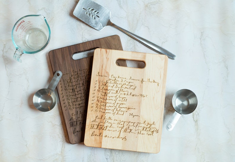Pode incluir: Duas t&aacute;buas de corte de madeira com receitas manuscritas, um copo medidor transparente, uma esp&aacute;tula de metal e uma x&iacute;cara de medi&ccedil;&atilde;o de metal. Uma t&aacute;bua &eacute; clara, a outra &eacute; escura. A esp&aacute;tula tem um desenho de folha.