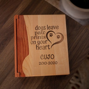 May include: A wooden pet memorial book with a light wood front and a darker wood spine. The front is engraved with the words "dogs leave paw prints on your heart" and a heart with paw prints. The name "CUJO" and dates "2010-2020" are also engraved.