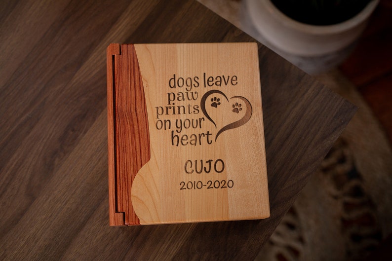 May include: A wooden pet memorial book with a light wood front and a darker wood spine. The front is engraved with the words "dogs leave paw prints on your heart" and a heart with paw prints. The name "CUJO" and dates "2010-2020" are also engraved.
