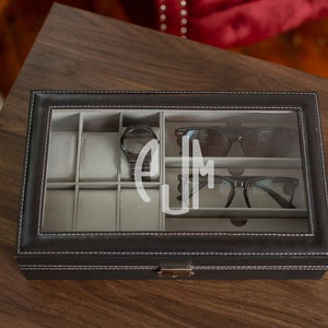 May include: A black leatherette watch and glasses organizer with a clear glass top. The interior has gray compartments for watches and eyewear. A black watch and two pairs of glasses are visible. The initials "EJM" are etched on the glass.