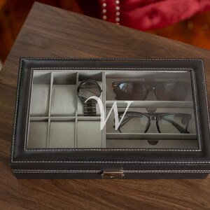 May include: Black leather watch and glasses organizer with a clear glass top. The organizer contains a black watch, sunglasses, and eyeglasses. A white letter "W" is visible on the glass. The organizer is on a wooden surface.