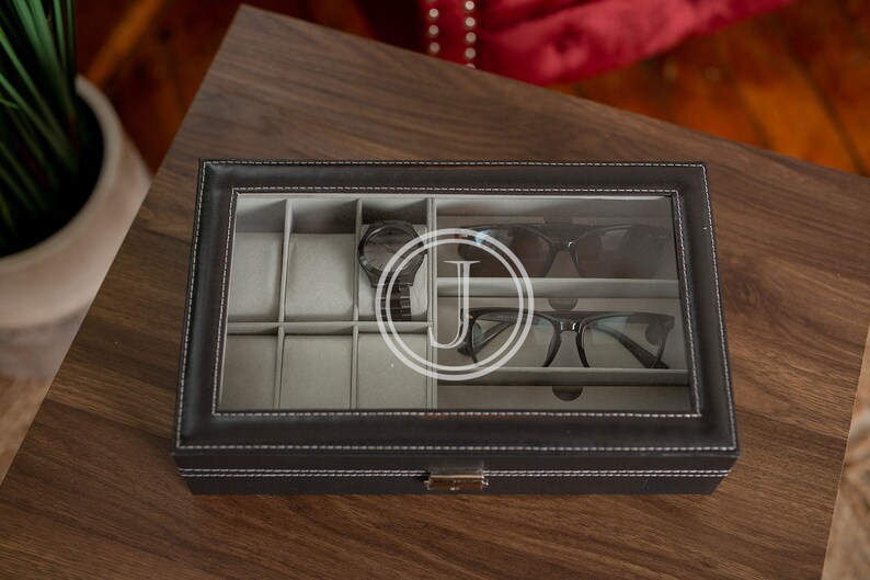 May include: Black leatherette watch and glasses organizer with a clear glass top. The organizer has multiple compartments for watches and eyewear. A watch, sunglasses, and eyeglasses are visible inside. The letter "J" is visible on the glass.