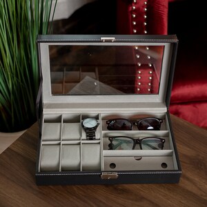 May include: A black leatherette watch and glasses organizer with a clear glass top. The interior has compartments for watches, sunglasses, and eyeglasses. A black watch and two pairs of glasses are inside the organizer.