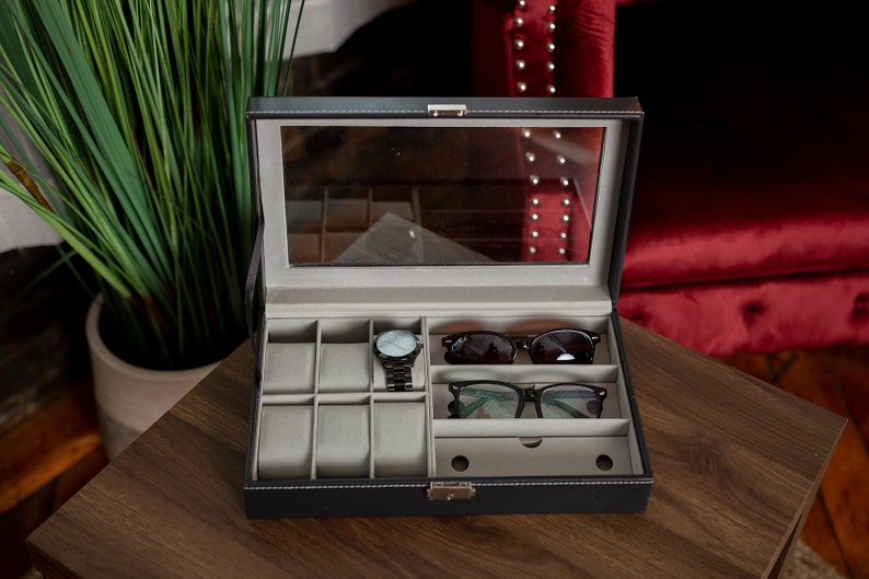 May include: A black leatherette watch and glasses organizer with a clear glass top. The interior has compartments for watches, sunglasses, and eyeglasses. A black watch and two pairs of glasses are inside the organizer.