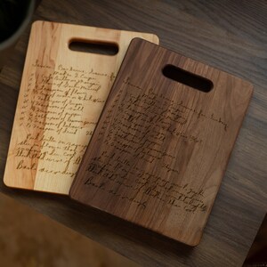 Pode incluir: Duas t&aacute;buas de corte de madeira, uma clara e outra escura, com formato retangular e uma al&ccedil;a. Cada t&aacute;bua tem uma receita manuscrita gravada em sua superf&iacute;cie. As t&aacute;buas s&atilde;o exibidas em uma mesa de madeira escura.