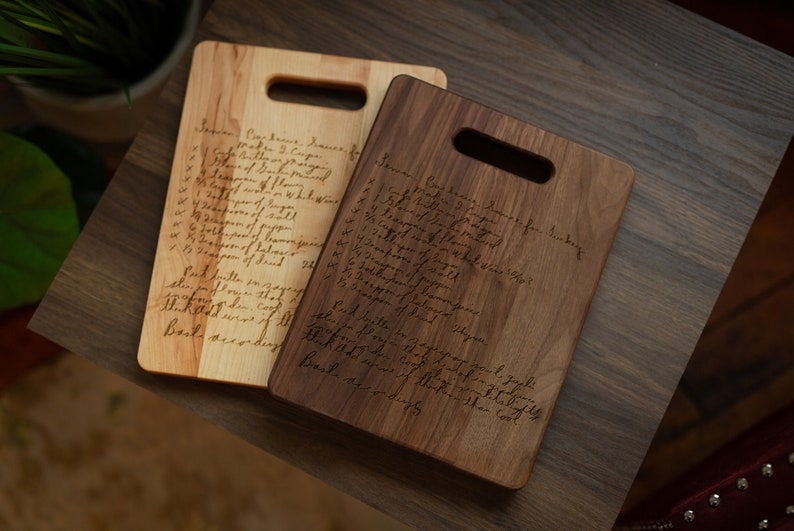 Pode incluir: Duas t&aacute;buas de corte de madeira, uma clara e outra escura, com formato retangular e uma al&ccedil;a. Cada t&aacute;bua tem uma receita manuscrita gravada em sua superf&iacute;cie. As t&aacute;buas s&atilde;o exibidas em uma mesa de madeira escura.