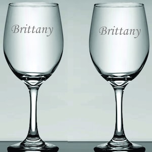 May include: Two clear wine glasses with a stem and bowl design. The name "Brittany" is etched on the side of each glass in a gray font. The glasses are displayed against a light gray background.
