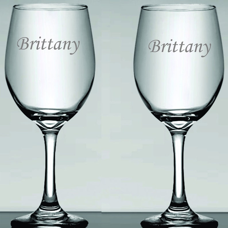 May include: Two clear wine glasses with a stem and bowl design. The name "Brittany" is etched on the side of each glass in a gray font. The glasses are displayed against a light gray background.