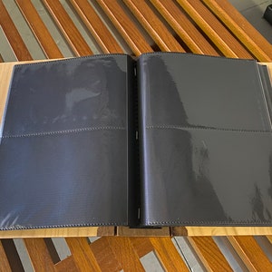 K&ouml;nnte beinhalten: Ein offenes schwarzes Fotoalbum mit transparenten Kunststoffh&uuml;llen, das auf einer hellen Holzoberfl&auml;che liegt. Das Album liegt auf einem Holztisch mit Lamellendesign. Das Album ist f&uuml;r Fotos konzipiert.