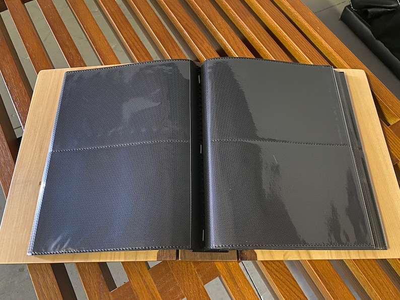 K&ouml;nnte beinhalten: Ein offenes schwarzes Fotoalbum mit transparenten Kunststoffh&uuml;llen, das auf einer hellen Holzoberfl&auml;che liegt. Das Album liegt auf einem Holztisch mit Lamellendesign. Das Album ist f&uuml;r Fotos konzipiert.