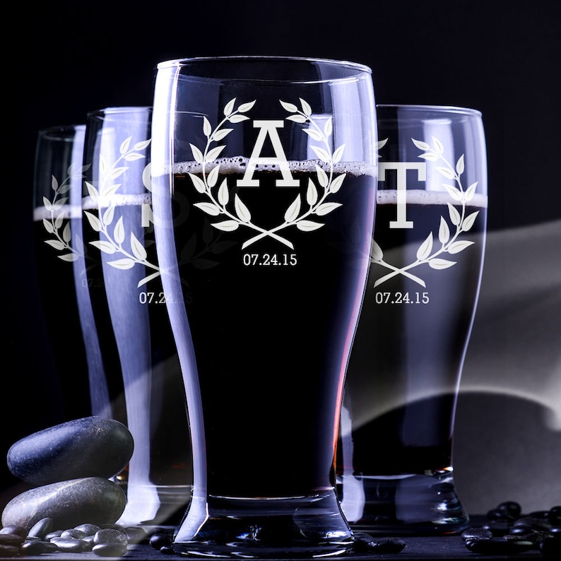 May include: Four tall, clear beer glasses filled with a dark liquid. Each glass is etched with a letter, a laurel wreath, and the date "07.24.15". The glasses are arranged in a row against a dark background, with some dark stones in the foreground.