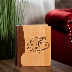 May include: A wooden box with a light wood front and a dark wood side. The front of the box has the words "dogs leave paw prints on your heart" with paw prints inside a heart shape. The box is sitting on a wooden surface.