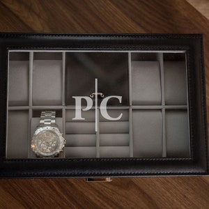 May include: Black leather watch box with a clear top, holding a silver wristwatch. The interior is gray with compartments for watches and rings. The initials "PC" are displayed on the back of the box.