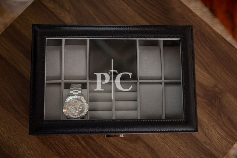 May include: Black leather watch box with a clear top, holding a silver wristwatch. The interior is gray with compartments for watches and rings. The initials "PC" are displayed on the back of the box.