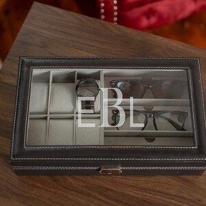 May include: Black leatherette watch and glasses organizer with a clear glass top. The interior is divided into compartments for watches, sunglasses, and eyeglasses. The initials "EBL" are visible on the glass. The organizer is on a wooden surface.