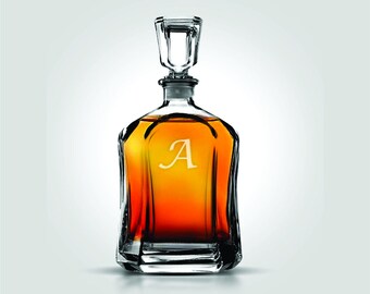 Peut inclure: Une carafe en verre transparent avec une base carrée et un bouchon. La carafe est remplie d'un liquide brun doré et porte la lettre "A" gravée sur le devant. Le bouchon est rectangulaire et transparent.