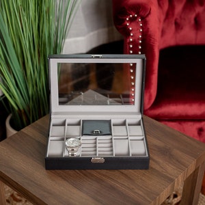 May include: Black watch and jewelry display case with a clear glass top, open on a wooden table. The interior is gray with compartments for watches and other accessories. A silver watch is inside the case.