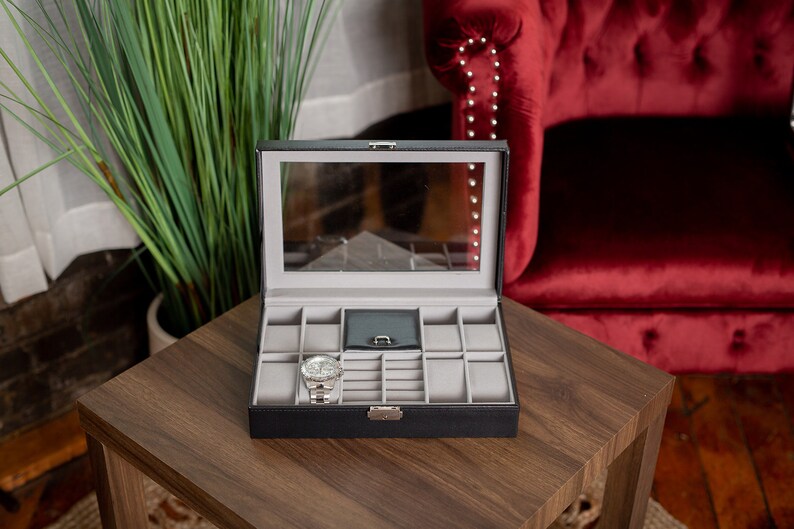 May include: Black watch and jewelry display case with a clear glass top, open on a wooden table. The interior is gray with compartments for watches and other accessories. A silver watch is inside the case.