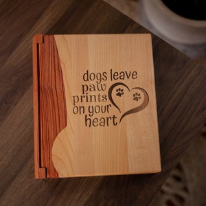 May include: A wooden photo album with a light wood front and a darker wood spine. The front is engraved with the words "dogs leave paw prints on your heart" and a heart with paw prints.