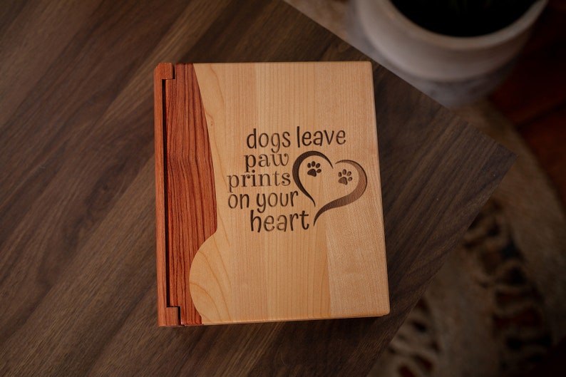 May include: A wooden photo album with a light wood front and a darker wood spine. The front is engraved with the words "dogs leave paw prints on your heart" and a heart with paw prints.