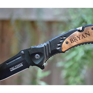 以下が含まれることがあります： 「BRYAN」という名前が刻まれた木製のハンドルが付いた、黒とシルバーの折りたたみナイフ。 刃は部分的に木の幹に差し込まれています。 ナイフの刃には「TAC-FORCE」の文字があります。