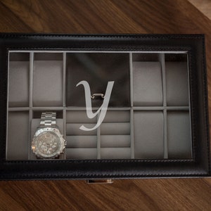 May include: Black leather watch box with a clear top, featuring a silver wristwatch. The interior is gray with compartments for watches and jewelry. A large, stylized letter "Y" is visible on the inside of the lid.