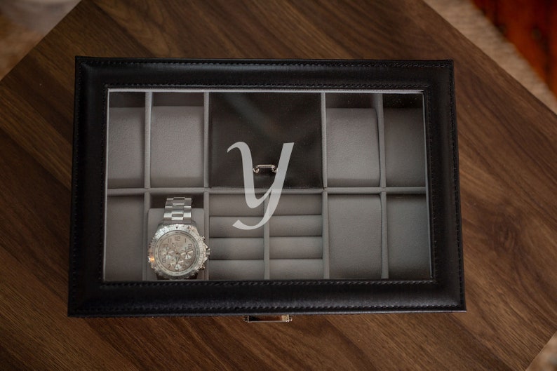 May include: Black leather watch box with a clear top, featuring a silver wristwatch. The interior is gray with compartments for watches and jewelry. A large, stylized letter "Y" is visible on the inside of the lid.