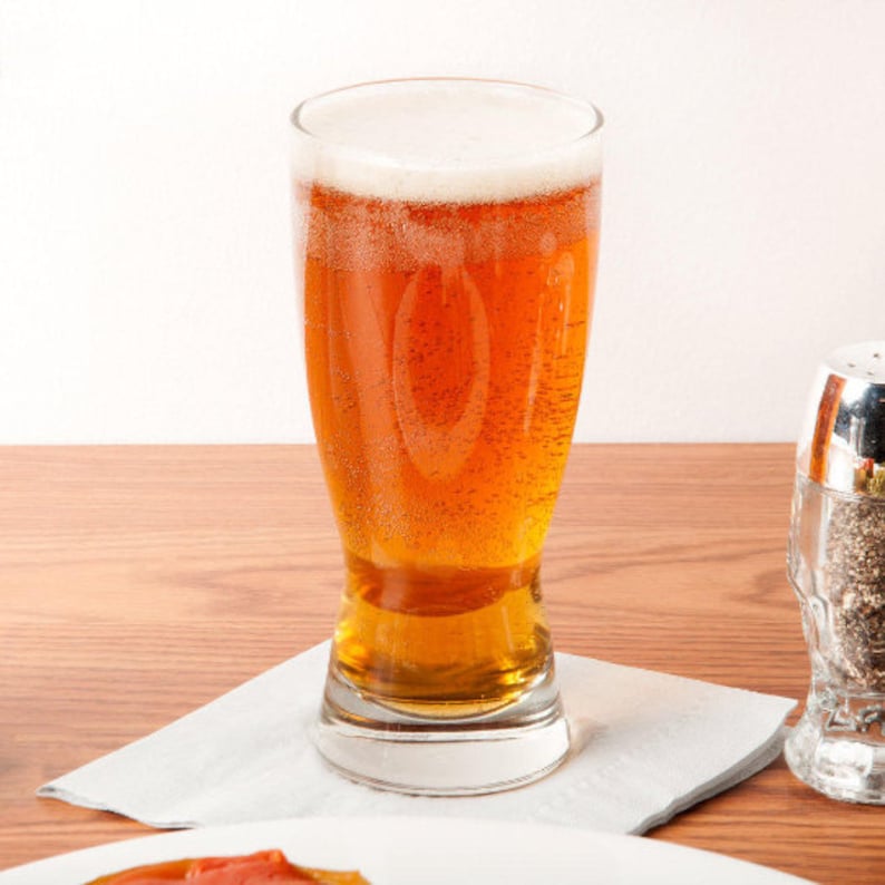 May include: A pint glass filled with amber beer and a white foamy head. The glass is on a white napkin, with a salt shaker in the background. The beer is bubbly and looks refreshing.