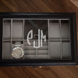 May include: Black leather watch box with a clear glass top, featuring a silver wristwatch and a monogram. The interior is lined with gray fabric and has compartments for watches and jewelry. The box is on a wooden surface.