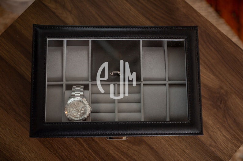 May include: Black leather watch box with a clear glass top, featuring a silver wristwatch and a monogram. The interior is lined with gray fabric and has compartments for watches and jewelry. The box is on a wooden surface.
