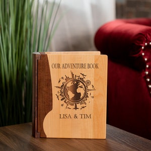 K&ouml;nnte beinhalten: Ein h&ouml;lzernes Abenteuerbuch mit einer hellen Holzfront und einem dunklen Holzr&uuml;cken. Die Vorderseite zeigt ein Weltkarten-Design mit dem Text "OUR ADVENTURE BOOK" und den Namen "LISA & TIM" darunter eingraviert. Das Buch steht auf einer Holzoberfl&auml;che.