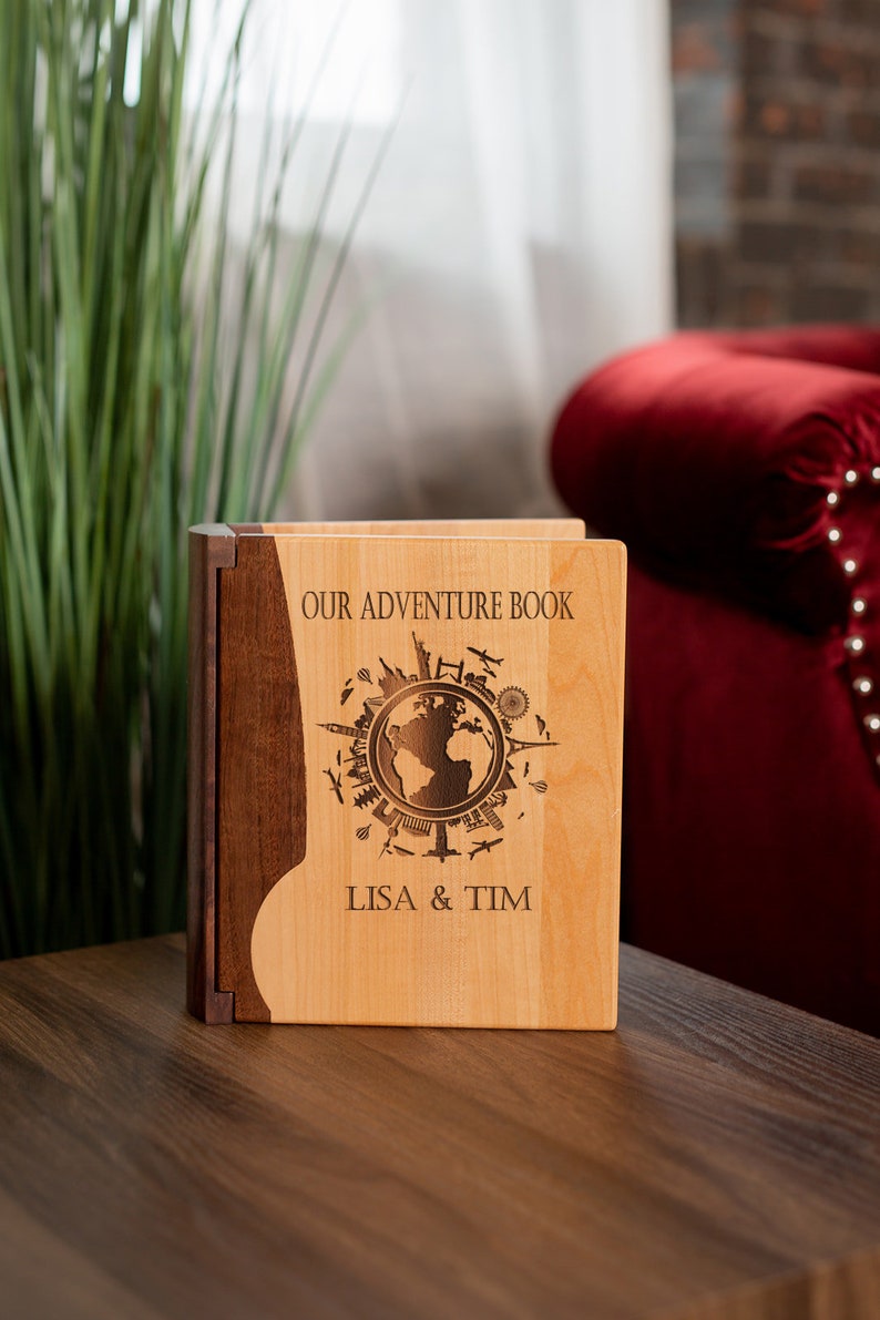 K&ouml;nnte beinhalten: Ein h&ouml;lzernes Abenteuerbuch mit einer hellen Holzfront und einem dunklen Holzr&uuml;cken. Die Vorderseite zeigt ein Weltkarten-Design mit dem Text "OUR ADVENTURE BOOK" und den Namen "LISA & TIM" darunter eingraviert. Das Buch steht auf einer Holzoberfl&auml;che.