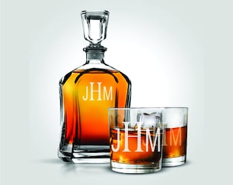 Pode incluir: Um decantador de vidro transparente e dois copos baixos a condizer, cada um gravado com as iniciais "JHM". O decantador está cheio de líquido âmbar. Um copo contém cubos de gelo. O conjunto foi concebido para servir bebidas espirituosas.