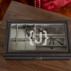 May include: Black leather watch and glasses organizer with a clear glass top. The interior has gray compartments for watches, sunglasses, and eyeglasses. The box is on a wooden surface.