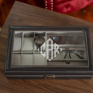 May include: Black leatherette watch and glasses organizer with a clear glass top. The interior has gray compartments for watches, sunglasses, and eyeglasses. A black watch, sunglasses, and eyeglasses are visible inside. The initials "CAR" are etched on the glass.