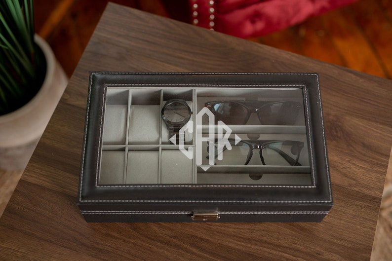 May include: Black leatherette watch and glasses organizer with a clear glass top. The interior has gray compartments for watches, sunglasses, and eyeglasses. A black watch, sunglasses, and eyeglasses are visible inside. The initials "CAR" are etched on the glass.