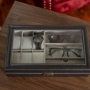 May include: A black leatherette watch and glasses organizer with a clear glass top. The interior has gray compartments for watches, sunglasses, and eyeglasses. A black watch and two pairs of glasses are visible inside the organizer. The organizer is on a wooden surface.