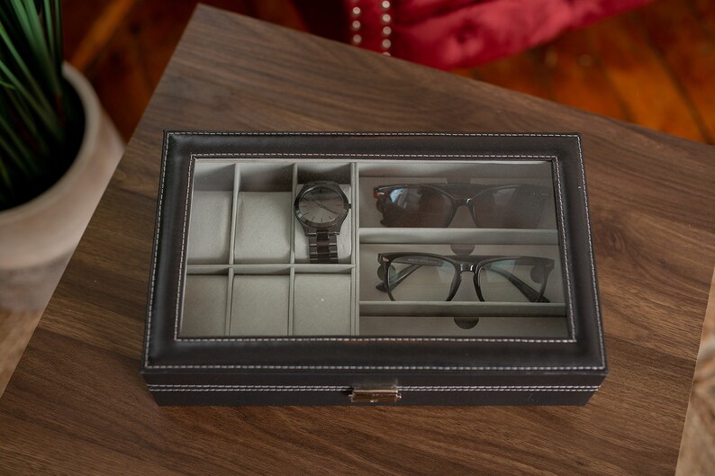 May include: A black leatherette watch and glasses organizer with a clear glass top. The interior has gray compartments for watches, sunglasses, and eyeglasses. A black watch and two pairs of glasses are visible inside the organizer. The organizer is on a wooden surface.