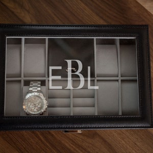 May include: Black leather watch box with a clear glass top, featuring gray velvet compartments for watches and jewelry. A silver wristwatch rests inside. The initials "EBL" are displayed on the back of the box.